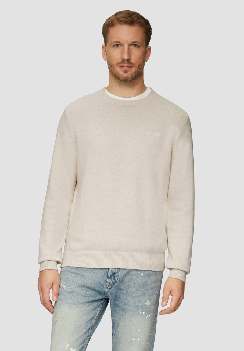 Man in een beige ronde hals trui over een wit shirt en lichtblauwe gescheurde jeans, staande tegen een effen lichte achtergrond.