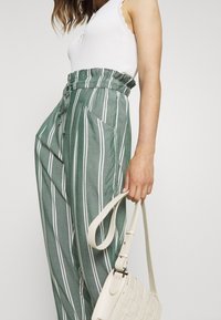 Pantalones a rayas verdes con una cinturilla elástica alta y bolsillos, combinados con una blusa blanca y llevando un bolso de mano tejido color crema.