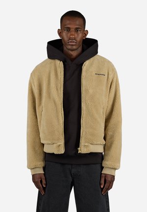 Beige Fleecejacke mit Reißverschluss, gerippten Bündchen und Saum. Über einem schwarzen Hoodie getragen, der ein kleines Logo auf der linken Brust hat.