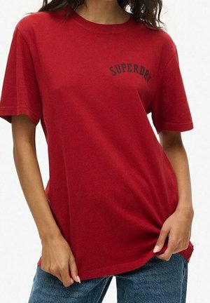 Henkilö pukeutuneena punaiseen Superdry T-paitaan ja sinisiin farkkuihin, pitäen paidan helmaa molemmilla käsillä.