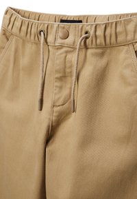 Pantalons kaki avec taille élastique, cordon de serrage, poches avant et fermeture à un bouton. Tissu lisse et texturé de couleur neutre.