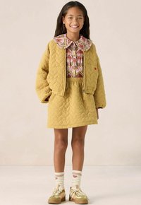 Veste matelassée jaune avec un col floral rose, jupe jaune assortie, chaussettes blanches rayées avec des cœurs, et baskets vertes.