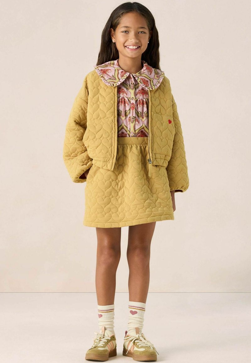 Veste matelassée jaune avec un col floral rose, jupe jaune assortie, chaussettes blanches rayées avec des cœurs, et baskets vertes.