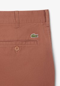 Pantaloni di cotone marroni con una finitura testurizzata, dotati di una tasca posteriore con bottone e un piccolo logo di coccodrillo verde e bianco sul lato destro.