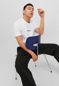 White cotton t-shirt with "Originals Studio" printed in black. Black trousers. Person sitting on a blue chair with a chrome accent.
