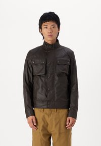 Σκούρο καφέ δερμάτινο μπουφάν με ψηλό κολάρο, δύο τσέπες στο στήθος και κουμπιά τυπου snap; εφαρμοστή γραμμή και λεία υφή. Φοριέται πάνω από μπεζ παντελόνι.