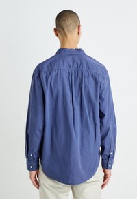 Camisa de manga larga azul marino con cuello abotonado, detalle de pliegues en la parte posterior y puños abotonados. Fabricada en suave tela de algodón.
