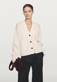 Crème ribbeltrui met een V-hals, knoopsluiting en voorzakken, gedragen over losse zwarte broeken. Houdt een bruine suède tas vast.