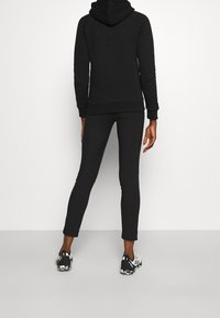 Schwarzer Hoodie mit strukturierter Oberflächenbeschaffenheit und eng anliegendem Schnitt, kombiniert mit schwarzen Leggings und sportlichen schwarz-weiß Sneakers, von hinten betrachtet.