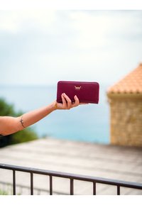 Burgundy leather wallet with a gold logo, zip closure, and a smooth texture, held in a hand against a blurred seascape background.