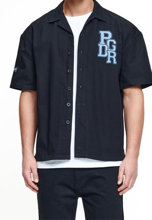 Mann trägt ein schwarzes kurzärmeliges Hemd mit blauen "PGDR" Buchstaben auf der Brust über einem weißen T-Shirt und schwarzen Hosen.