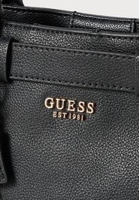 Černá kožená kabelka s texturou, představující zlaté logo "GUESS" a nápis "EST 1981", se strukturálním designem a viditelným švem.