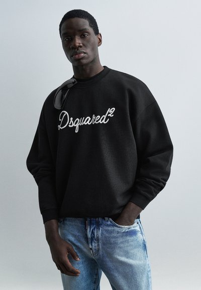 Νέος άνδρας που φοράει μαύρο φούτερ Dsquared² και ανοιχτόχρωμο τζιν, με γυαλιά ηλίου να κρέμονται από τη λαιμόκοψη, στέκεται μπροστά σε μονόχρωμο γκρι φόντο.