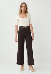 Roomkleurige top met vierkante hals, gecombineerd met hoge, donkere, breed-uitlopende broek, op de taille aangetrokken met een bruine riem en afgewerkt met naakte hoge hakken.
