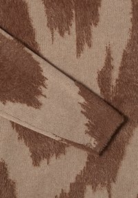 Close-up van beige en bruine donzige gebreide trui stof met abstracte, onregelmatige patroonen zichtbare mouwdetails.
