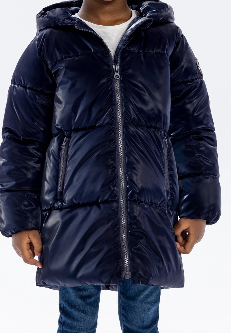 Enfant portant un manteau d'hiver long matelassé bleu marine avec une fermeture éclair frontale et des poches latérales zippées, associé à un jean bleu.