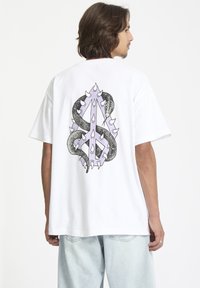 T-shirt en coton blanc à manches courtes, présentant un design graphique d'un serpent s'entrelacant avec un symbole de paix épineux au dos.