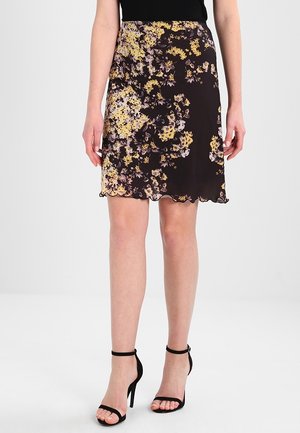 Femme portant une jupe noire à fleurs jaunes et violettes, assortie à des sandales noires à talons hauts avec des brides à la cheville.