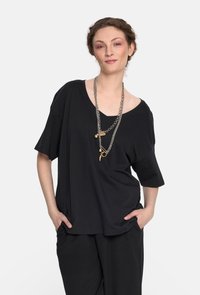 Czarny, oversize krótki top z szerokim dekoltem, wykonany z miękkiego materiału. Połączony z czarnymi, luźnymi spodniami i warstwą srebrnych naszyjników.