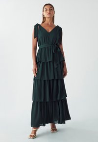Grünes, schichtweise gestaltetes Maxikleid mit V-Ausschnitt, verstellbaren Schulterbändern und einem Taillengürtel. Leichter Stoff mit fließender Silhouette.