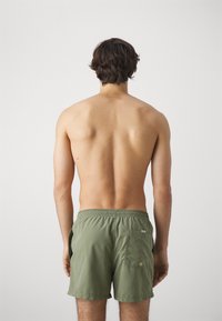 Olijfgroene zwembroek met een elastische tailleband, achterzak en oogletdetails. Het beeld toont de achterkant en onthult een minimalistisch ontwerp.