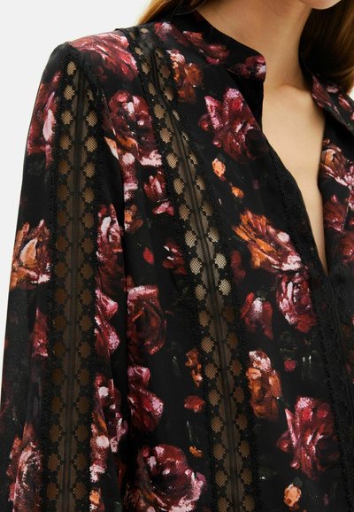 Blusa floreale nera con motivi di rose rosse e arancioni. Presenta dettagli in pizzo lungo le maniche e un design senza colletto. Tessuto leggero.