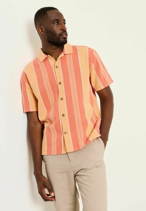 Mann trägt ein kurzärmliges Hemd mit orange-gelben vertikalen Streifen, beigen Hosen, blickt zur Seite und hat eine Hand in der Tasche.