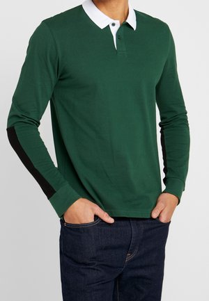 Homme portant un polo à manches longues vert foncé avec col blanc et coudières noires, mains dans les poches d'un jean bleu foncé.