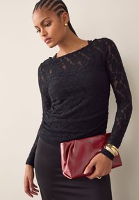 Zwart kanten top met lange mouwen en een ronde halslijn met een gestructureerd patroon, gecombineerd met een rode leren clutch en gouden accessoires, tegen een lichte achtergrond.