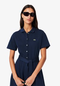 Abito polo blu navy con maniche corte, chiusura a bottoni sul davanti e cintura annodata in vita. Presenta un piccolo logo verde Lacoste. Indossato con occhiali da sole scuri.