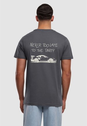 Mann trägt graues T-Shirt mit weißer Sportwagen-Illustration und dem Text „Never too late to the party“ auf dem Rücken, kombiniert mit hellblauen Jeans.