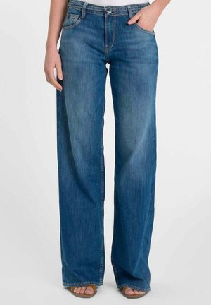 Vrouw die een hoge taille, wijd uitlopende blauwe spijkerbroek draagt met steekzakken aan de voorkant, een knoopsluiting en open-toe sandalen, staand tegen een effen achtergrond.