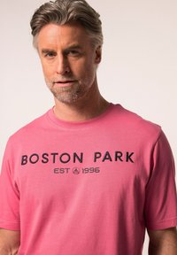 Růžové bavlněné tričko s černým vyšitým textem "BOSTON PARK EST 1996." Krátké rukávy a kulatý výstřih s hladkou texturou.