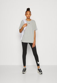 Camiseta gris de Nike con logo negro, combinada con leggings negros que tienen un logo blanco. Modelo sosteniendo una mochila lavanda con zapatillas deportivas.