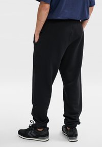 Schwarze Jogginghose mit elastischem Bund und Bündchen, ausgestattet mit seitlichen Taschen und lockerer Passform. Getragen mit schwarzen Sportschuhen.