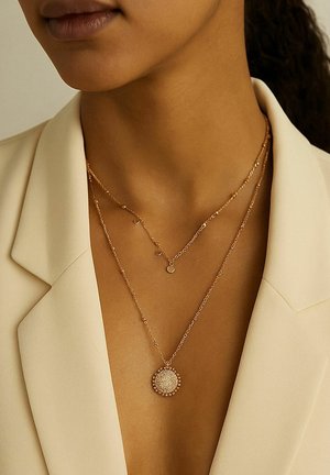 Schichtweise angeordnete Halsketten aus Roségold mit einem kreisförmigen Anhänger mit strukturierten Oberflächen und kleinen Perlen. Getragen über einem hellen Blazer.