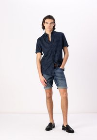 Chemise à manches courtes marine avec des pois blancs, assortie à un short en denim roulé. Le modèle porte des mocassins noirs, debout contre un fond neutre.