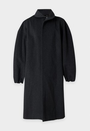 Homme portant un manteau sombre oversize, une chemise grise, un pantalon gris et des chaussures noires, debout devant un fond clair uni.