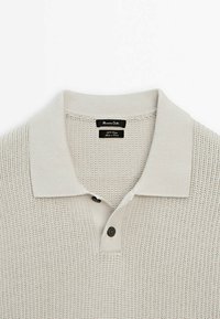 Vaaleanharmaa neulottu poolopaita, jossa on teksturoitu kudos, lyhyet hihat ja klassinen kaulus. Siinä on kaksi tumman sävyistä nappia ja kudottu merkki.