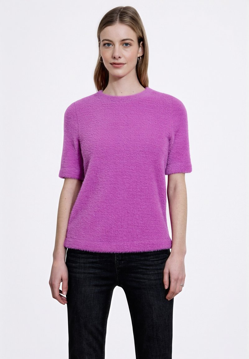 Chlupatý růžový svetr s krátkým rukávem a kulatým výstřihem, kombinovaný s tmavými džíny. Má měkkou texturu a rovný lem.
