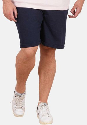 Homme portant un short bleu marine à revers, des baskets blanches à lacets et une chemise de couleur claire, marchant vers l'avant sur un fond uni.