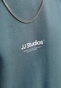 Groene katoenen t-shirt met witte bedrukte logo "JJ Studios" en een zilveren ketting, gemaakt van een gladde, gestructureerde stof.