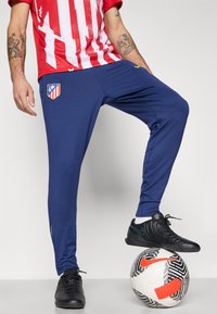 Sportbukser i navy blå med en slim fit, med et klublogo-patch, parret med sorte sportssko på en mønstret fodbold.