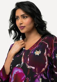 V-Ausschnitt Bluse mit floralen Mustern auf schwarzem Grund, die lebendige lila und gelbe Blumen zeigt, aus weichem Stoff gefertigt und mit drei Viertelärmeln.