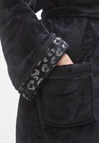 Albornoz de forro polar negro que presenta una superficie texturizada, un detalle de estampado de leopardo en los puños y un bolsillo frontal. Material suave y cálido.