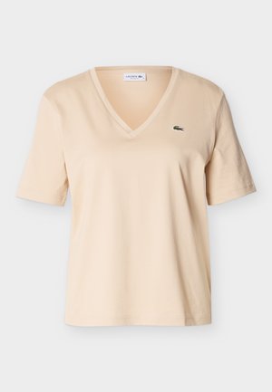 Μπεζ T-shirt με V-λαιμόκοψη και κοντά μανίκια, με μικρό πράσινο λογότυπο κροκόδειλου στο αριστερό στήθος, εμφανιζόμενο σε απλό φόντο.