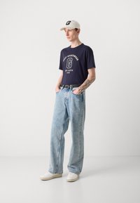 Marineblå t-skjorte med hvit logo, lyseblå løse jeans, svart belte og hvite joggesko. Tilbehøret inkluderer en beige baseball caps.