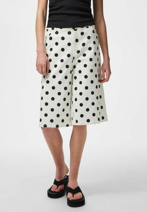 Osoba nosiaca biele široké šortky s čiernymi bodkami, čierne tielko bez rukávov a čierne platformové sandále.