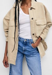 Veste beige oversized avec grandes poches, fermeture à boutons et coupe décontractée, portée sur un débardeur blanc et un jean bleu clair.
