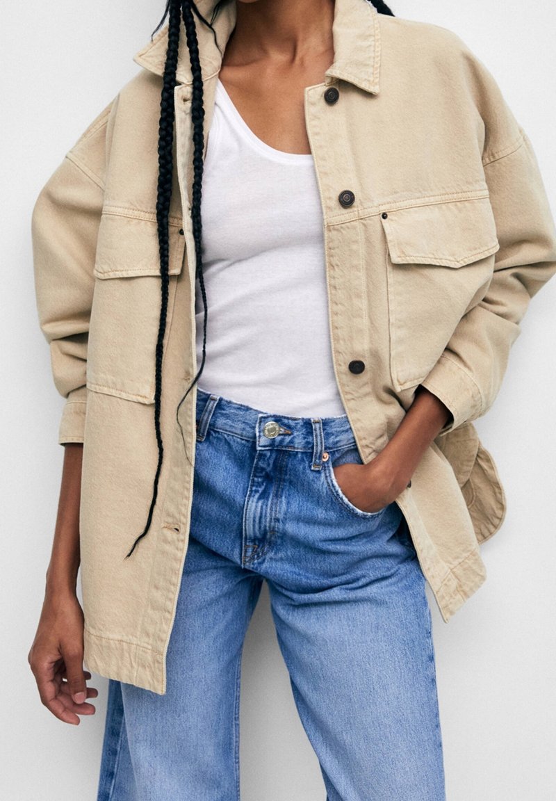 Veste beige oversized avec grandes poches, fermeture à boutons et coupe décontractée, portée sur un débardeur blanc et un jean bleu clair.